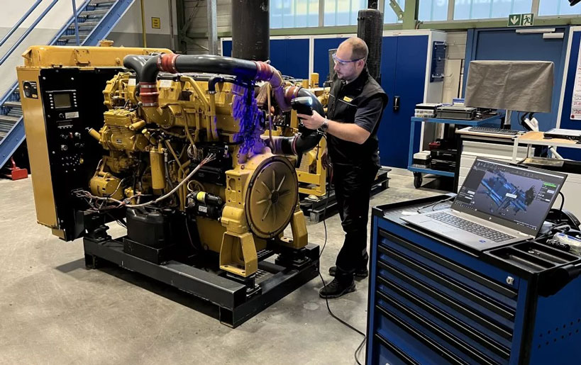 Man in a black polo shirt with yellow Zeppelin logo and black trousers scanning a yellow-colored engine block with the HandySCAN BLACK 3D scanner. On the right in the foreground is a laptop on a blue work trolley on which the scan can be seen