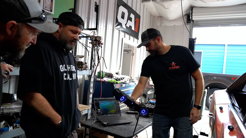 Three technicians looking at a computer screen as one of them 3D scans a monster truck.