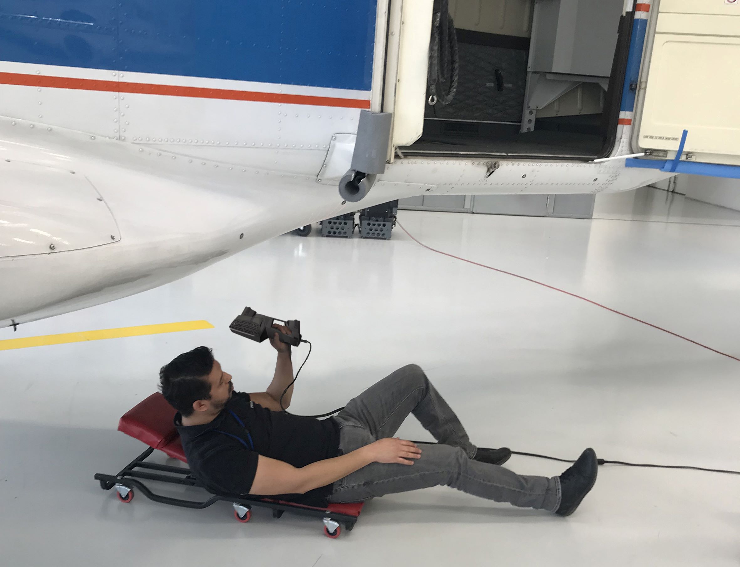 Technician on a mechanic creeper using the HandySCAN EVO Elite scanner to scan below a white, blue and red plane.