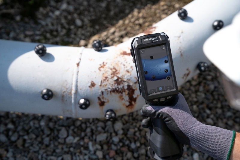 HandySCAN EVO|Elite handheld 3D scanner capturing data and displaying the scan on its built-in screen while inspecting a rusted white oil pipe with multiple black markers attached.