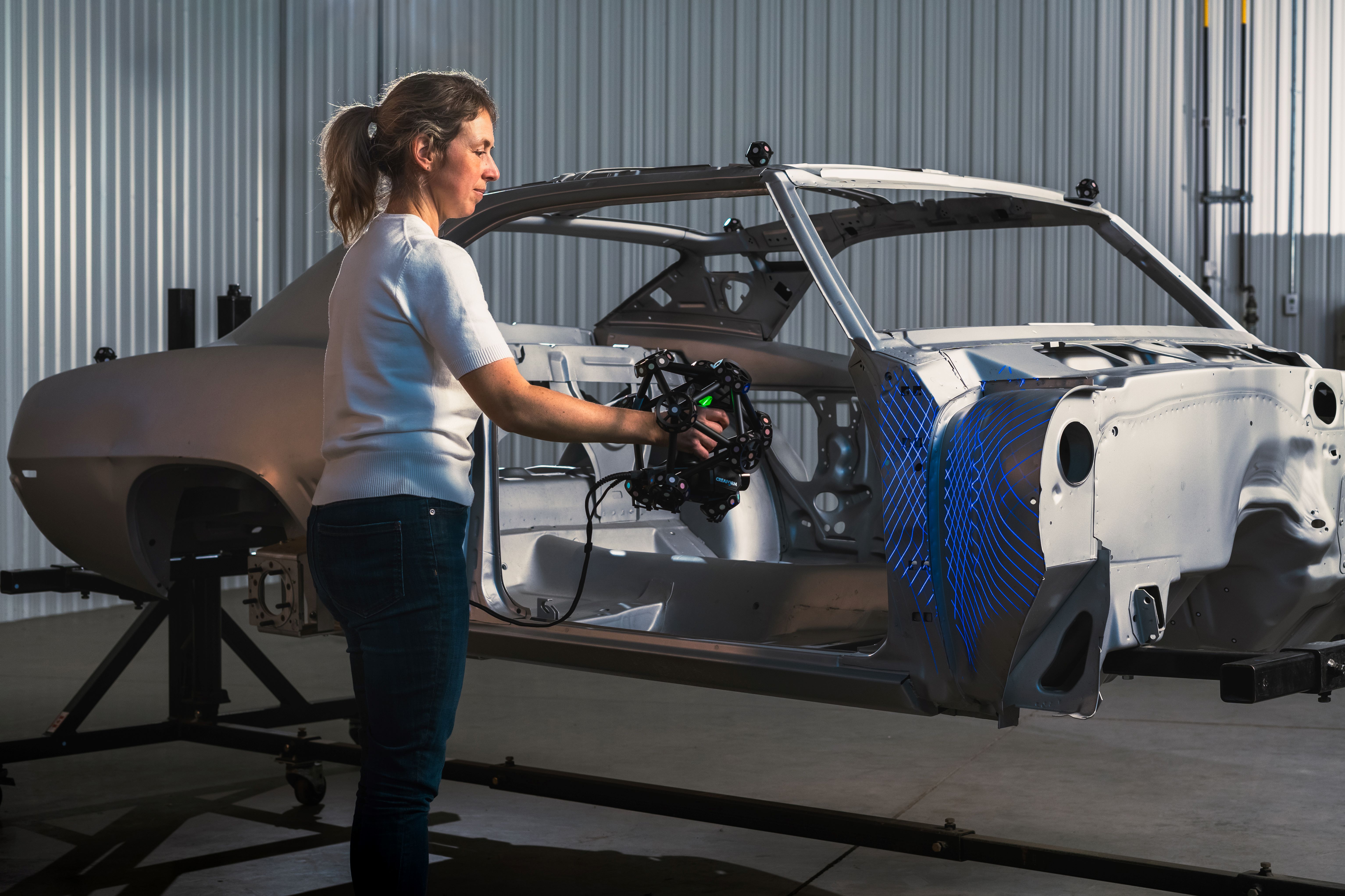 Una donna in un magazzino che utilizza MetraSCAN 3D per misurare un telaio grigio di un’auto montato su un telaio di supporto.