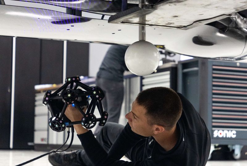 Engineer from WTR team lying on the floor using a Creaform MetraSCAN 3D scanner to capture the underside of a Cadillac WTR V-Series GTP race car, with live mesh projection visible on the surface.