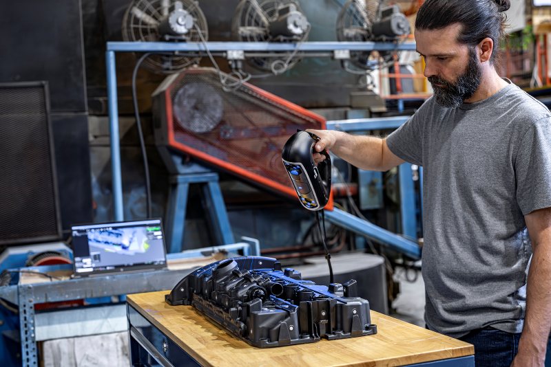 Engineer using a HandySCAN  3D to scan a black automotive intake manifold, with the 3D model displayed on a nearby computer screen.