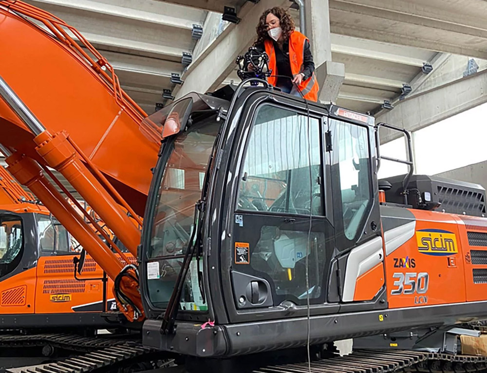 Woman with curly hair and a mouth mask scanning the roof of an excavator cab with the MetraSCAN 3D. The driver's cab consists mostly of glass panes, the excavator is orange