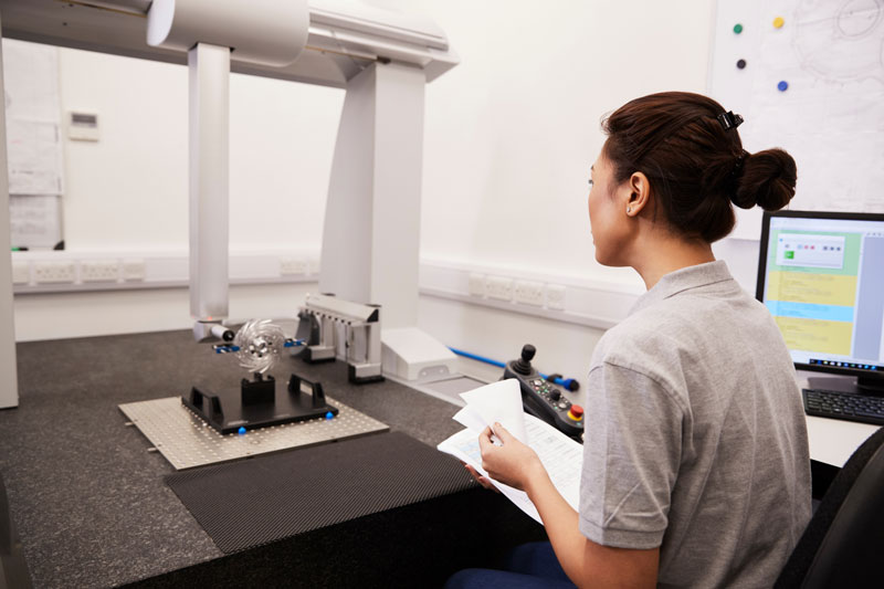 Woman employee sitting in front of a bridge CMM measuring a small aluminum circular part while sitting next to a computer screen