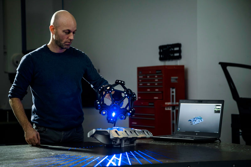 Funcionário usando um escâner 3D MetraSCAN para medir um coletor de admissão V8 em uma mesa ao lado de um laptop que exibe a peça digitalizada no Creaform.OS