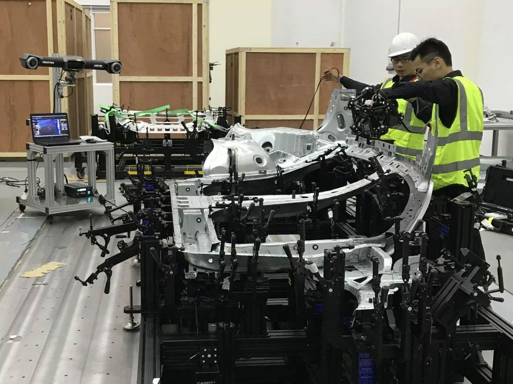 Two employees wearing yellow safety vests in a workshop using the MetraSCAN 3D|BLACK to measure a large stamped metal car part in a jig setup in front of the C-track and a laptop displaying the scanned data while another part is in a separate jig in the background
