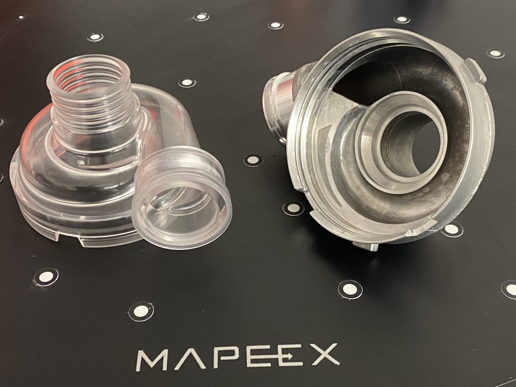 Transparentes Modell und Original Wasserpumpengehäuse liegen nebeneinander auf einer Unterlage, die mit Targets beklebt ist. Das Mapeex Logo ist zu sehen.
