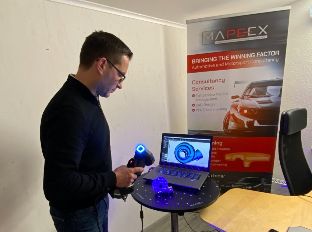 Ein Mann scannt ein Bauteil. Es sind blaue Laserlinien zu sehen. Der Scan ist auf dem Laptop zu sehen. Im Hintergrund steht ein Mapeex Banner.