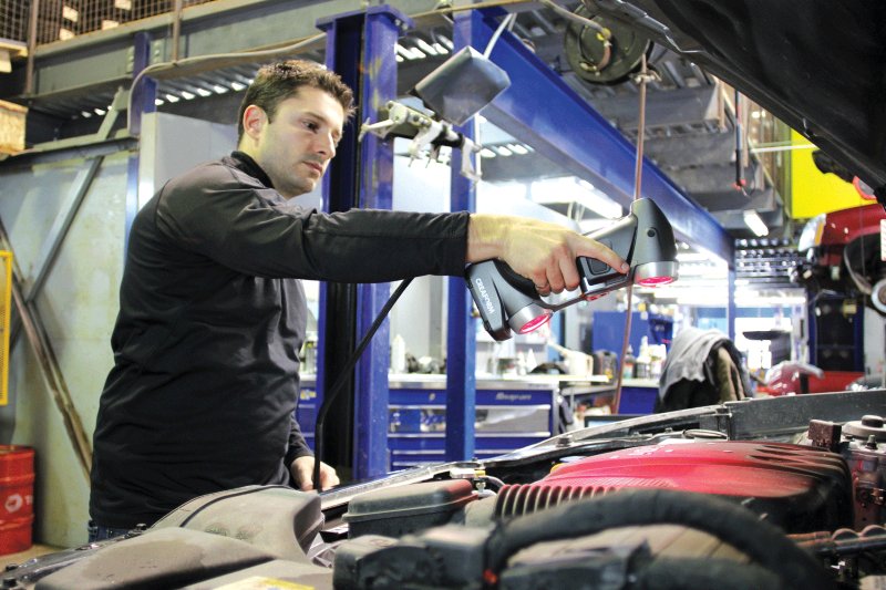 Personne utilisant un scanner 3D HandySCAN 3D portable sur le moteur d’un véhicule au sein d’un atelier industriel, avec des outils et des machines en arrière-plan