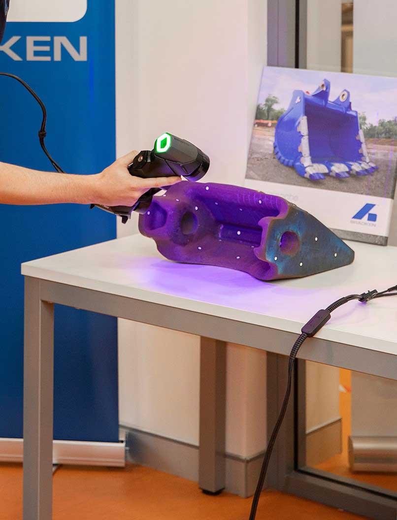 Un trabajador de Bradken utiliza el escáner HandySCAN 3D para escanear el diente de un cangilón de excavadora sobre una mesa frente a la imagen de un cangilón azul.
