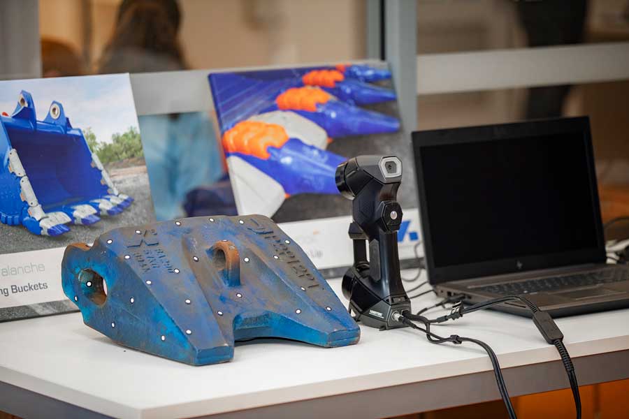 El escáner HandySCAN 3D se encuentra sobre una mesa, entre un diente azul del cangilón de la excavadora y una computadora portátil negra in front of pictures of the product.