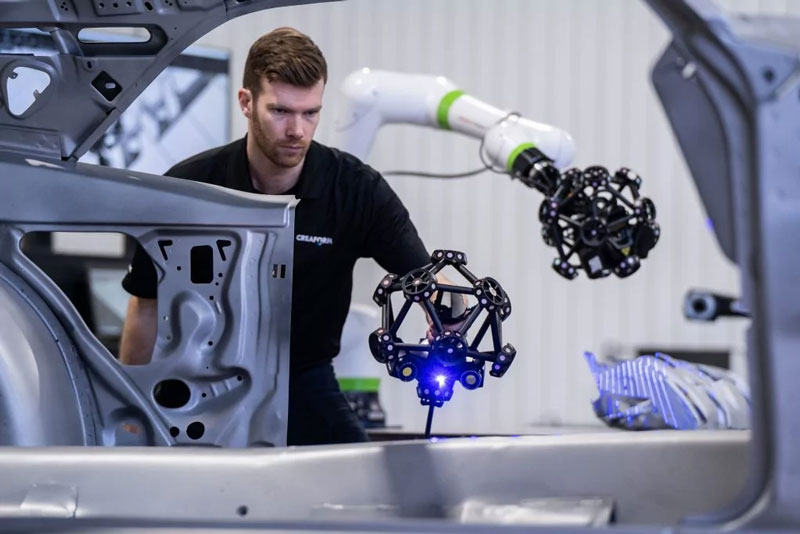 Employee using a MetraSCAN 3D scanner to measure a car frame next to a MetraSCAN 3D-R mounted on a cobot