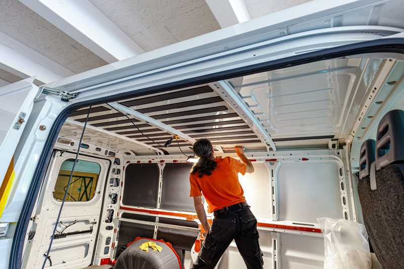 Hombre con pelo largo y playera naranja preparando el interior de una furgoneta blanca para su personalización.