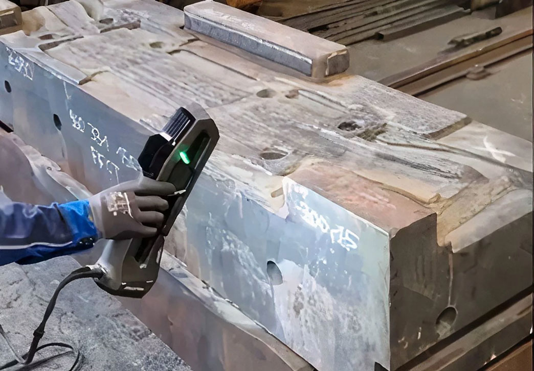 From the left: A hand with a gray glove holds a Go!SCAN 3D scanner with which a forging die for a truck axle is scanned