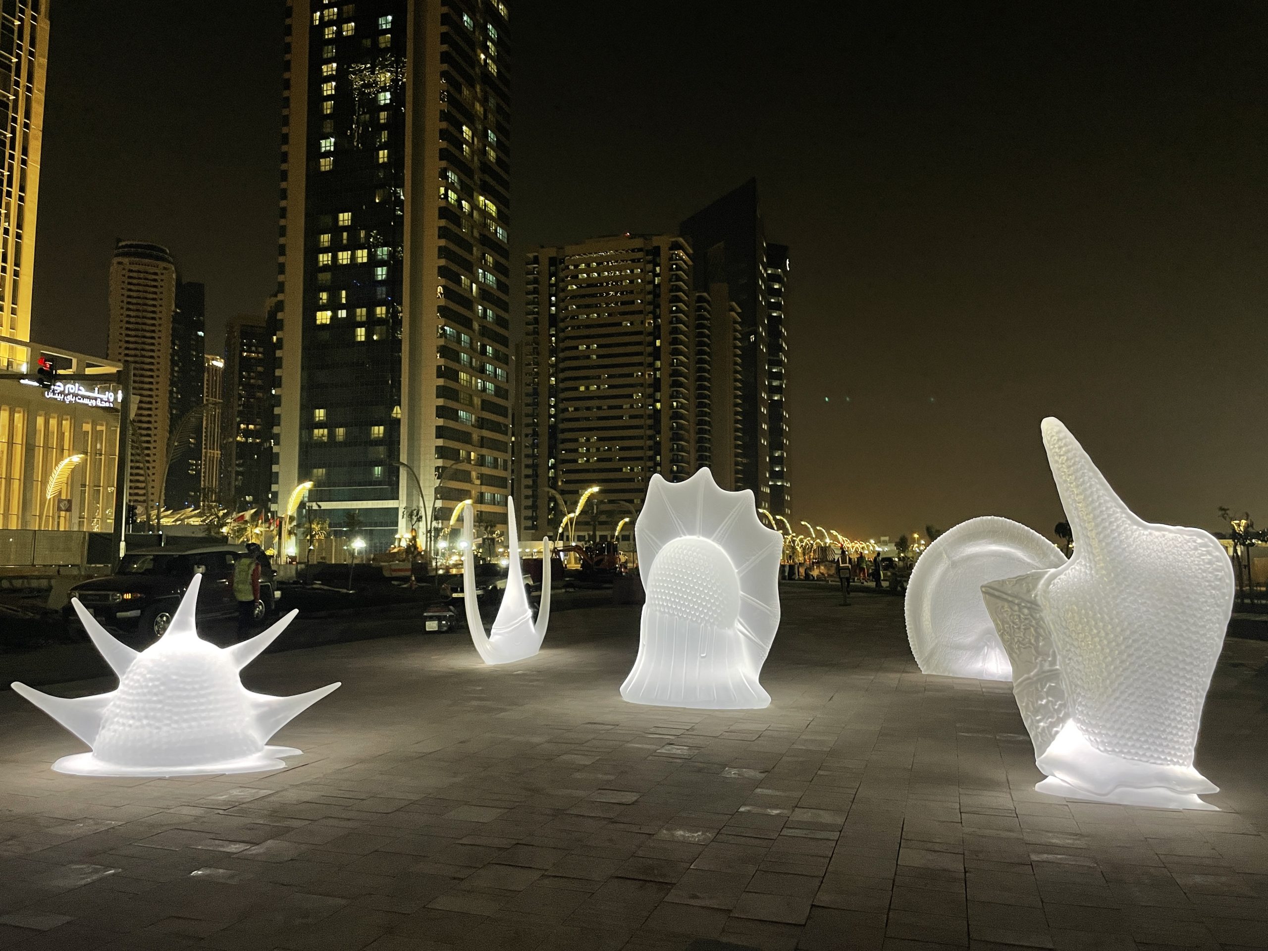 Sculture illuminate di notte. Le sculture sono immerse nella luce bianca. In secondo piano ci sono dei grattacieli. Alcune finestre sono illuminate.