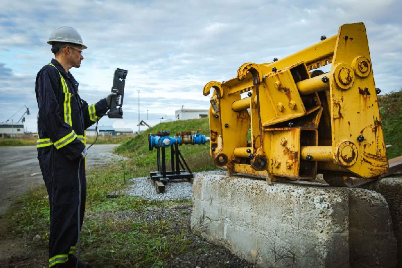 Man in safety jumpsuit wearing a hard hat using a HandySCAN 3D MAX Series scanner outside to scan a forklift attachment on a concrete block.