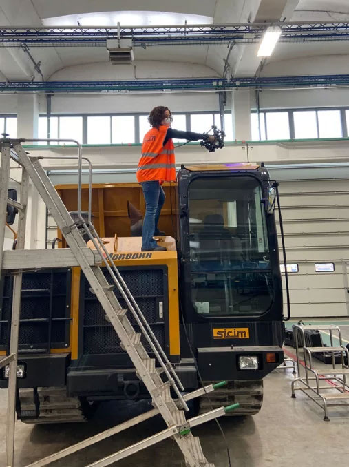 Woman standing on a yellow dump truck using a MetraSCAN 3D scanner to measure the driver cabin inside a warehouse.