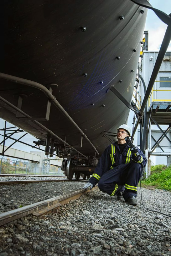 Employee wearing a one-piece worker suit and a hard hat under a tank-car to measure it with the HandySCAN 3D|MAX Series scanner, directly on the rails