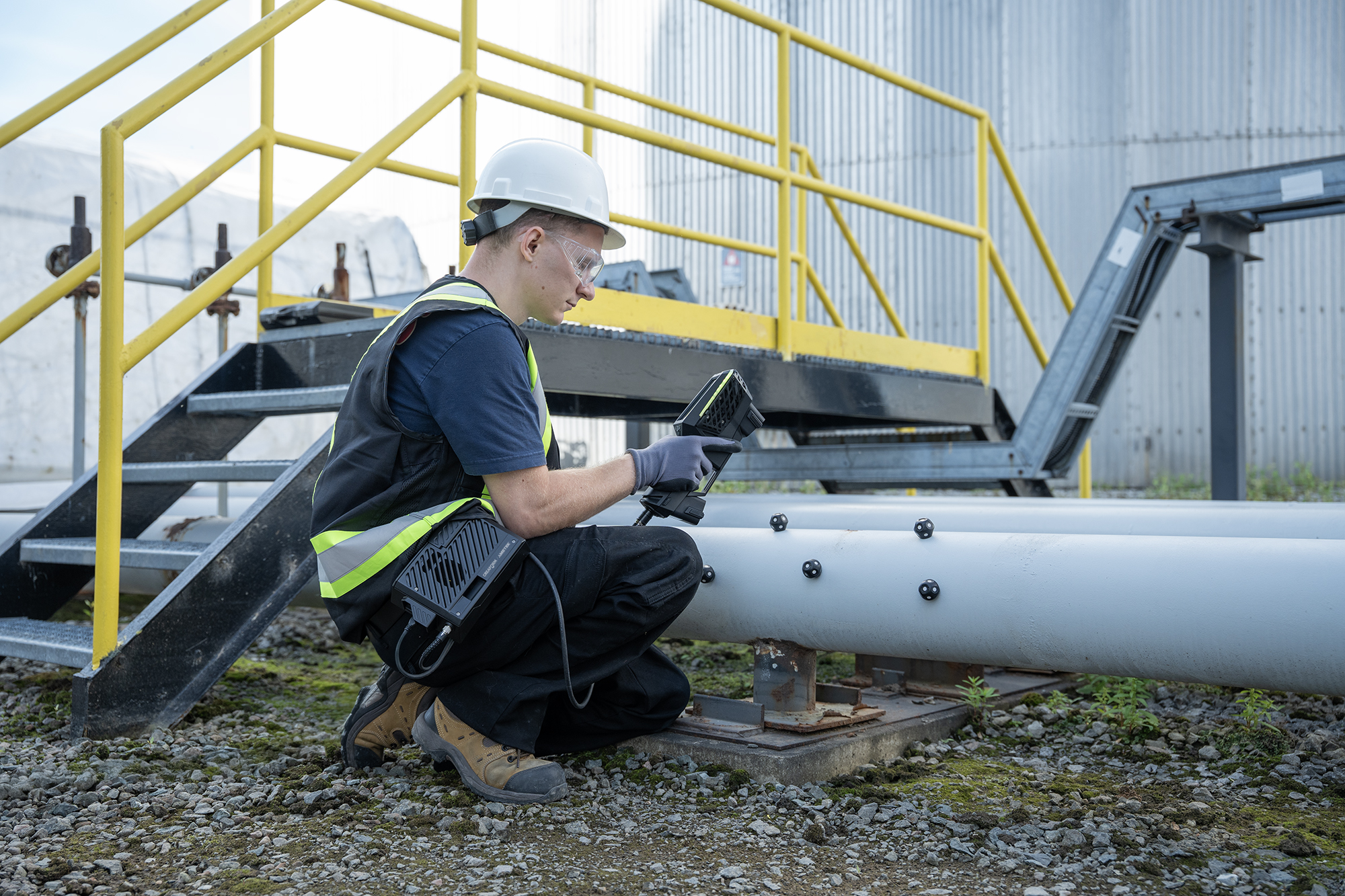 A technician wearing a hard hat and a safety vest crouching next to crossover stairs while scanning refinery pipes with the HandySCAN EVO Elite with the Mobility Kit to scan.