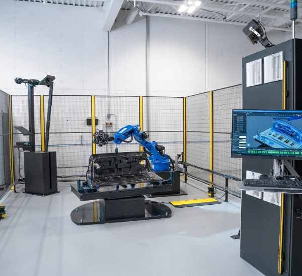 Industrial cell with blue robot holding a MetraSCAN 3D-R scanning a black sheet metal part, monitor shows 3D model