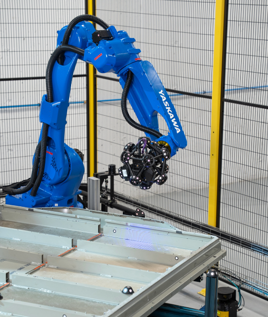 Blue Yaskawa robotic arm with MetraSCAN 3D scanner inspecting a large metallic panel inside safety enclosure