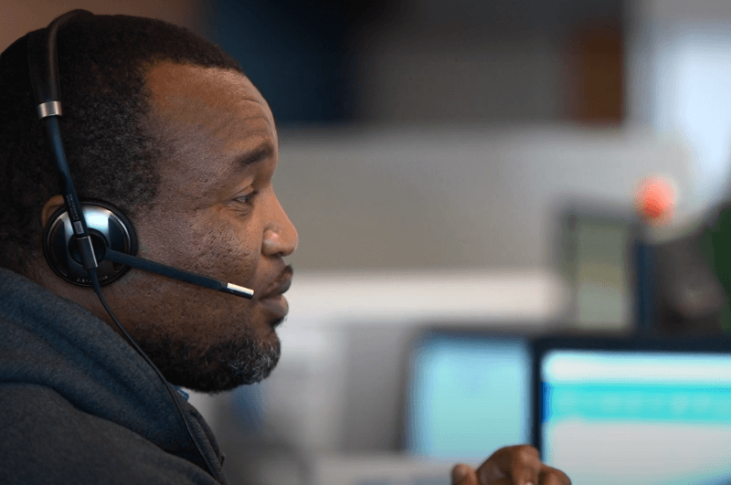 Customer service representative wearing a headset, seated at a desk with computer screens in a professional office environment.