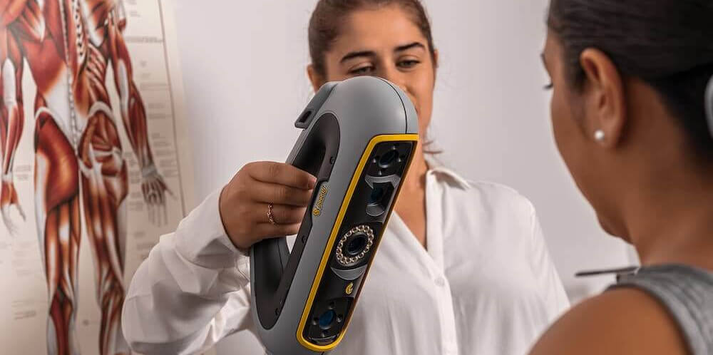 Healthcare professional using a Creaform Go!SCAN 3D scanner to capture a 3D image of a patient's body, with an anatomy chart visible in the background.