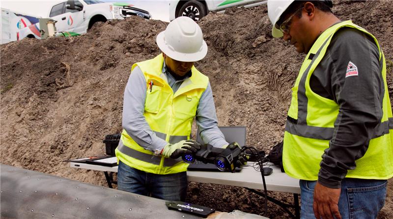 Two Integridad de Ductos employees in a ditch next to a pipeline and a table with a laptop using the HandySCAN BLACK|Elite to asses the integrity of the pipeline with two pickups in the background