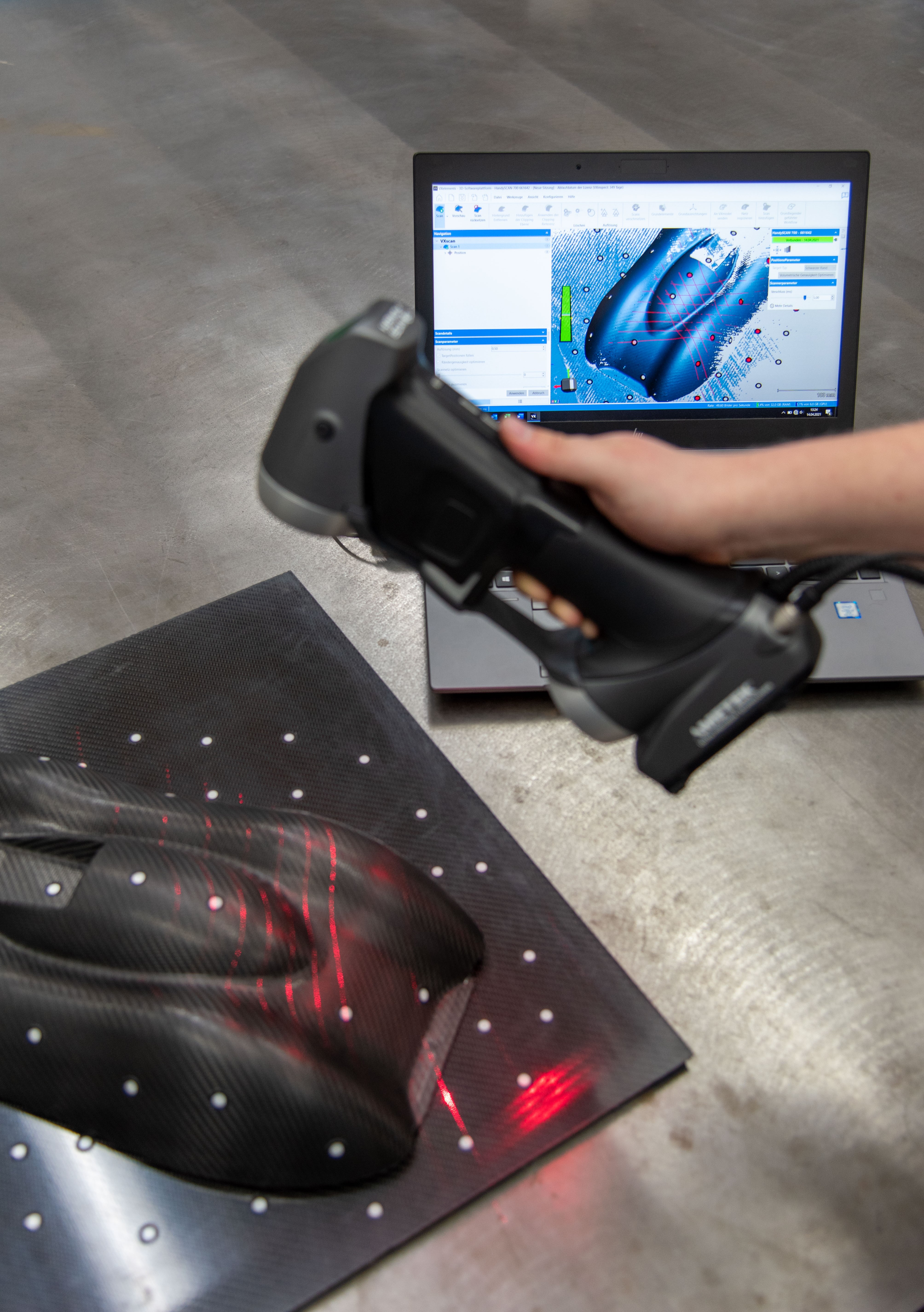 Employee using HandySCAN 3D SILVER series scanner to scan a carbon fber car part on a table in front of laptop displaying in real-time in VXelements
