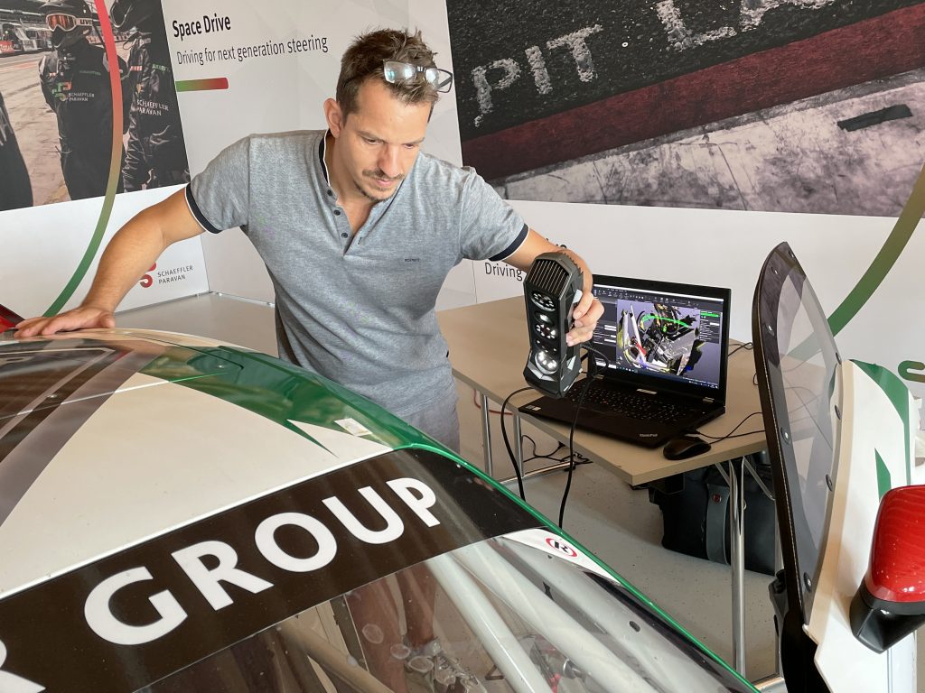 Homme avec une chemise grise utilisant le scanner Go!SCAN 3D pour numériser l'intérieur latéral d'une Porsche Cayman GT4 blanche, avec un ordinateur portable en arrière-plan affichant VXscan