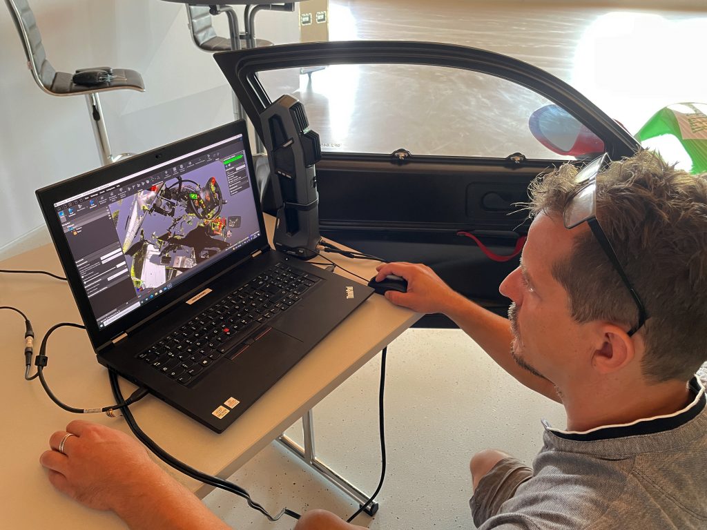穿灰色衬衫的男子在笔记本电脑前，笔记本电脑显示的是 VXelements 软件平台中白色保时捷 Cayman GT4 的内部扫描结果