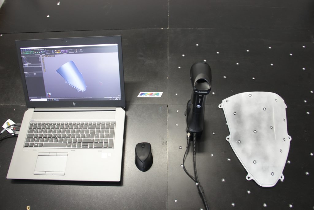 HandySCAN 307 放在一张桌子上，旁边是一块经过喷粉处理的摩托车挡风玻璃和一台屏幕上显示蓝色 3D 模型的笔记本电脑