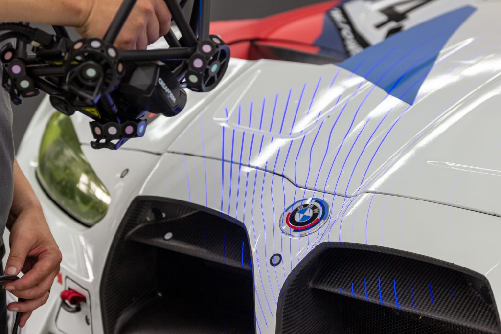 El MetraSCAN 3D escanea el capó de un motor con el logotipo de BMW utilizando líneas láser azules.