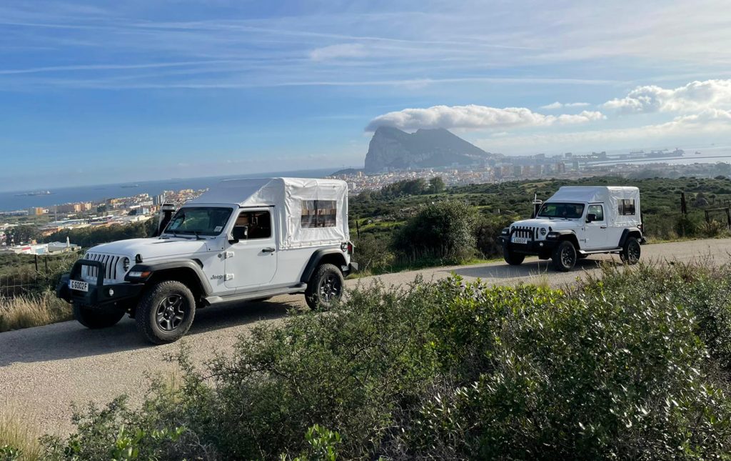 Two white jeeps on a road. In the background you can see the sea with ships, the rock of Gibraltar and a city.