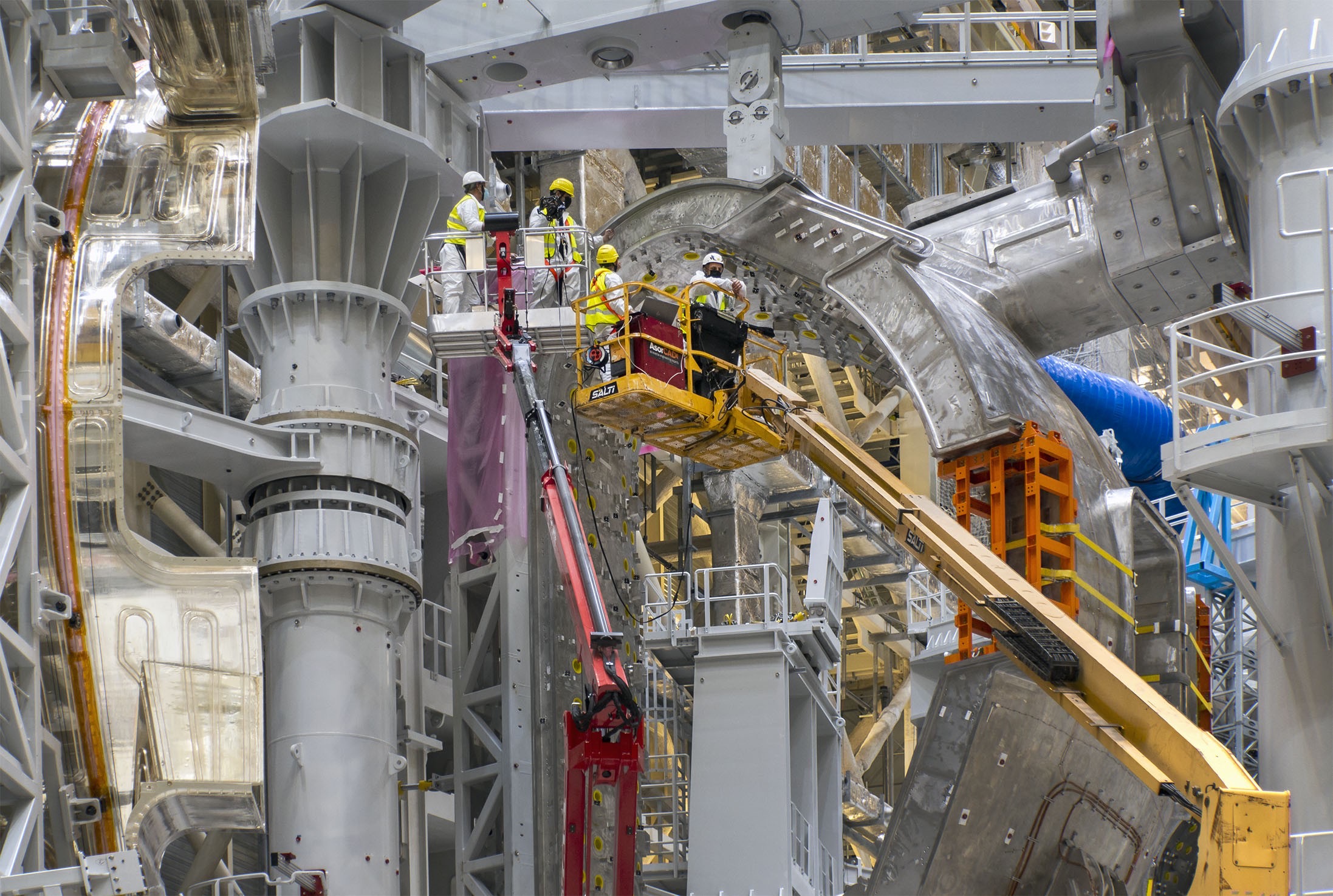 Wide view of the working environment of the ITER project, with two extended cranes with platforms on which four people with helmets and safety vests are standing.