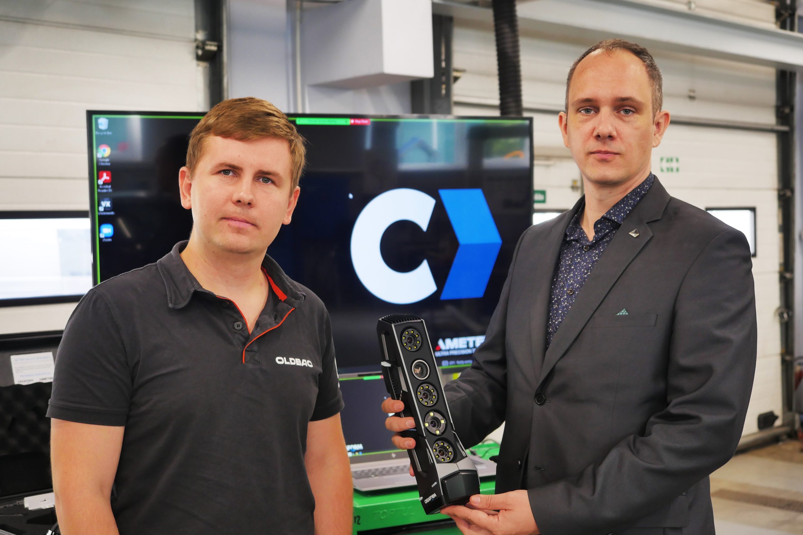 Two men in front of a TV screen which shows the Creaform logo icon. One of the man helds a Go!SCAN 3d scanner in his right hand.