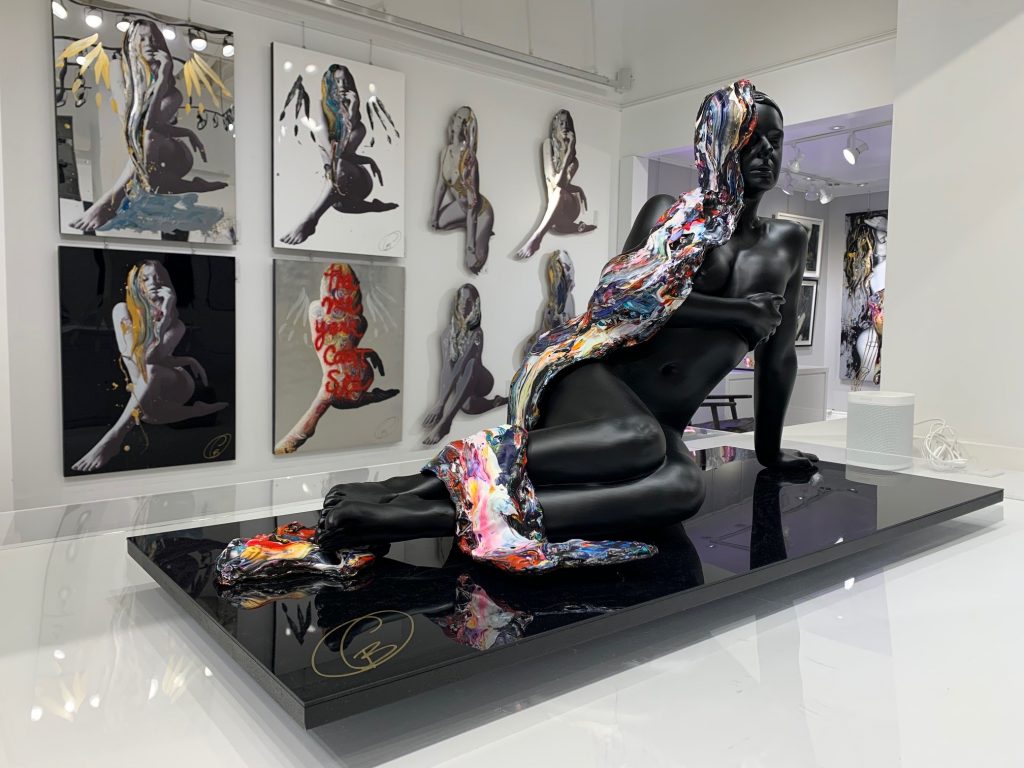 A finished black sculpture with colored hair on a table in the art gallery in front of several paintings