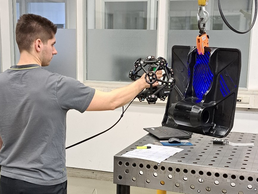 A team member uses the MetraSCAN 3D scanner to scan a black part that is suspended from a fixture above a metal table with holes. The blue laser beams of the scanner help to improve accuracy, reliability and repeatability. 