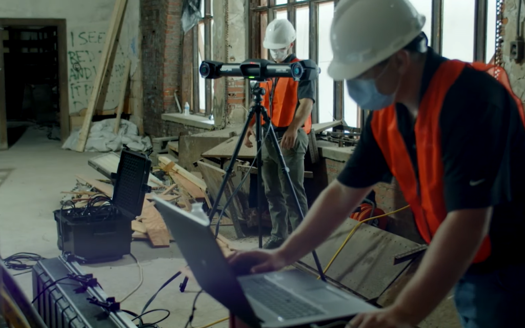 在废墟中的一个房间里，两名头戴白色安全帽的工人在 C-Track 旁的笔记本电脑上工作