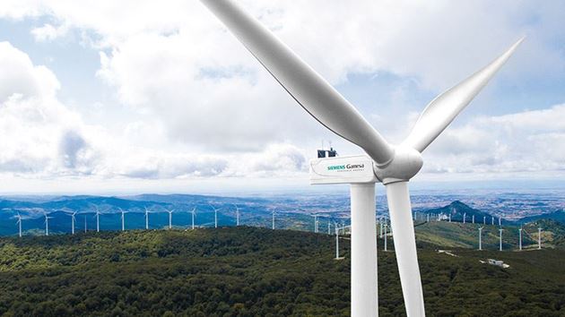 En primer plano un aerogenerador con el logo de Siemens Gamesa delante de una paisaje montañoso boscoso con más turbinas eólicas.