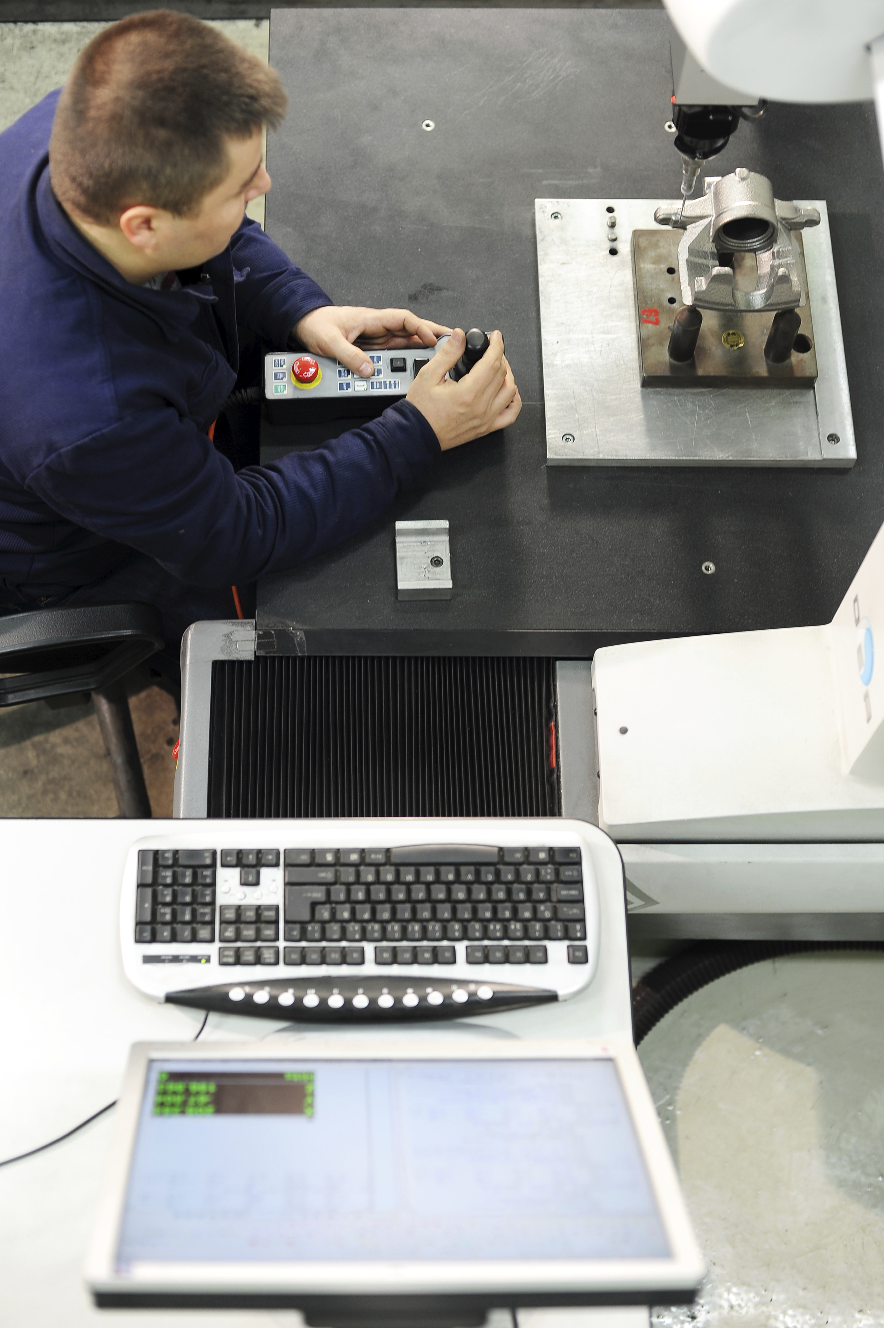 Vista superior del hombre midiendo una pieza metálica pequeña con CMM tradicional de contacto junto a la computadora