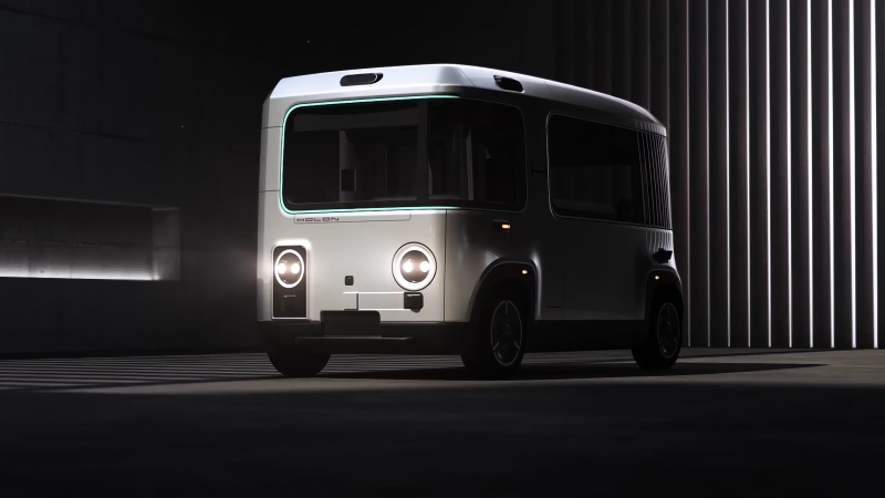 Transporte urbano eléctrico autónomo HOLON exhibido en un entorno arquitectónico moderno, con faros LED circulares y un diseño minimalista y aerodinámico.