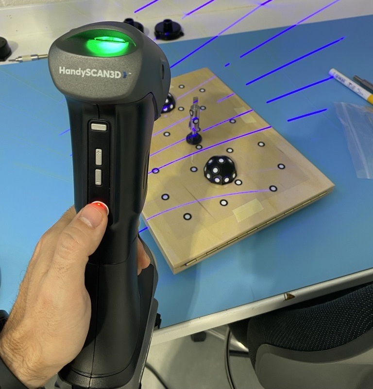 Close-up of a HandySCAN 3D handheld scanner projecting blue laser lines to digitize a small aerospace component.